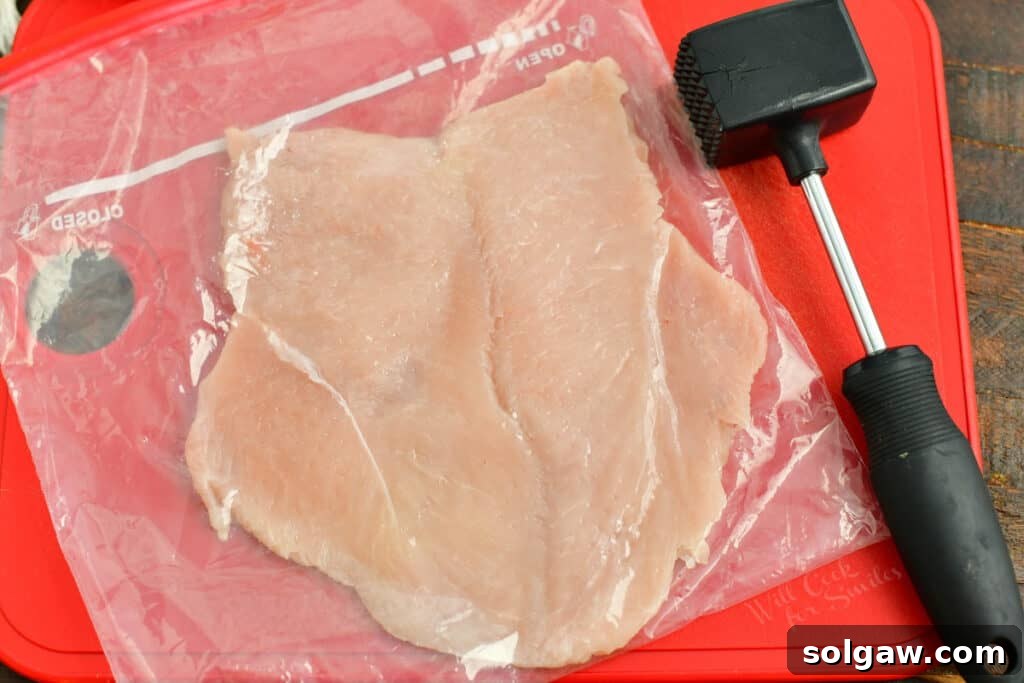 A flattened chicken breast on a red cutting board, with a meat mallet nearby, illustrating the pounding process.