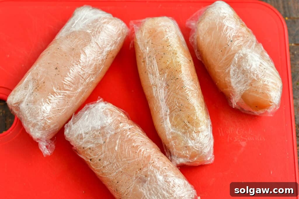 Chicken cutlets expertly rolled into cylinder shapes and wrapped tightly in plastic wrap, prepared for chilling.