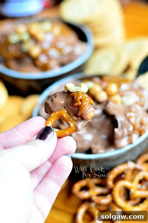 dipping a pretzel into Chocolate Fruit Dip in a blue bowl with pretzels to the right