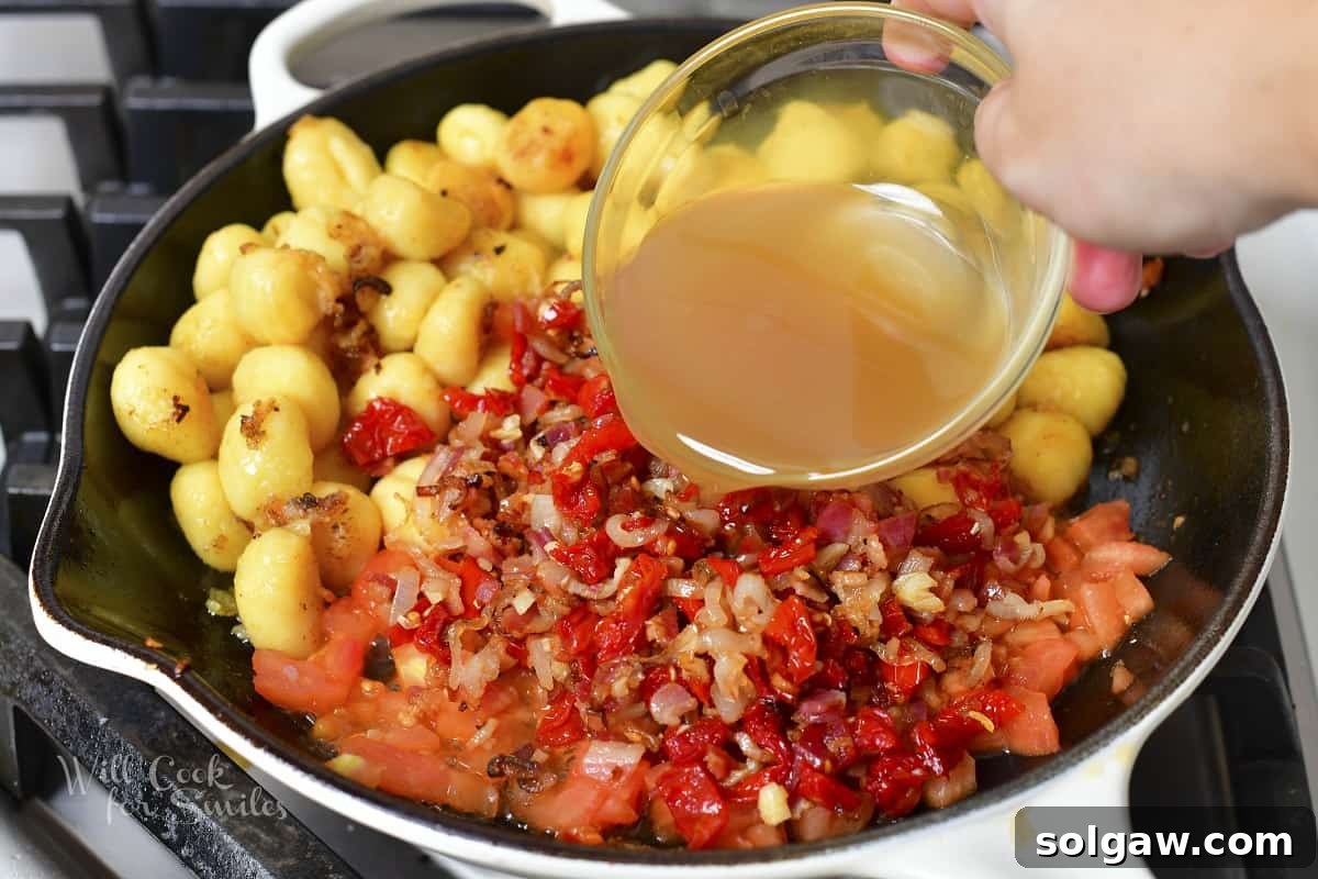 Pouring chicken stock into the pan with gnocchi and tomatoes, creating the base for the creamy sauce.