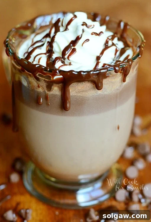 Hot Chocolate in a glass with whipped cream and chocolate sauce on a wood table with chocolate chips around it