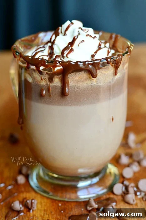 Hot Chocolate in a glass with whipped cream and chocolate sauce on a wood table with chocolate chips around it
