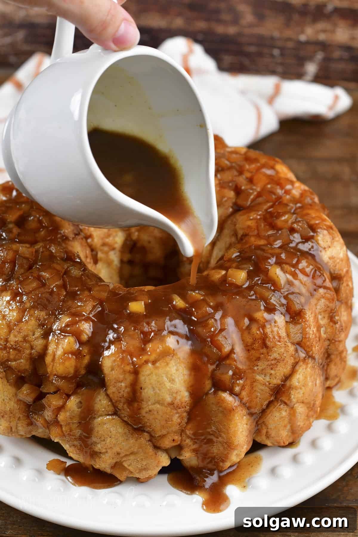 Pouring extra apple sauce over the freshly baked apple pie monkey bread on a white serving plate.