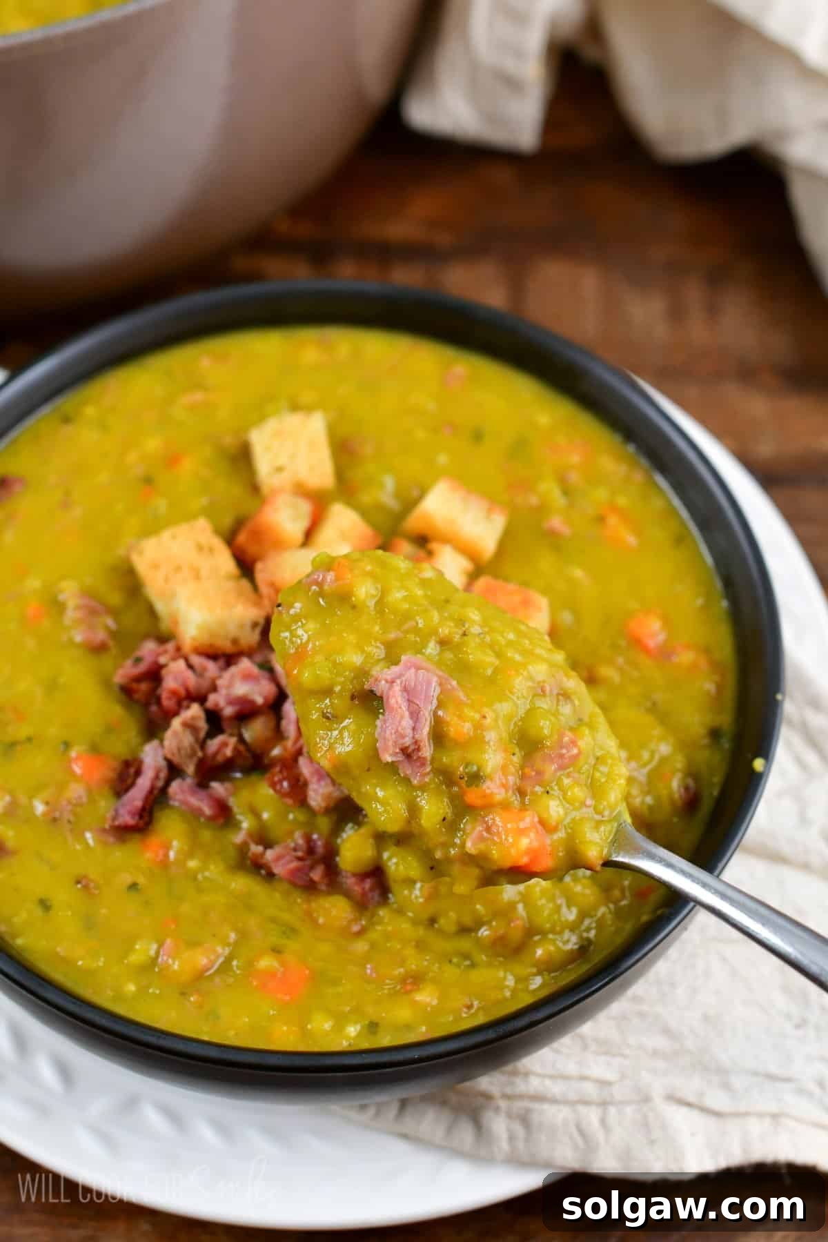 A generous serving of split pea soup in a bowl, topped with diced ham and croutons, with a spoon scooping a portion, highlighting its thick and satisfying texture.