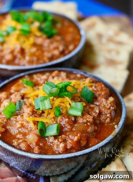 Two small bowls of rich, boozy chili, garnished with shredded cheese and fresh chives, artfully arranged on a blue platter with scattered crackers.
