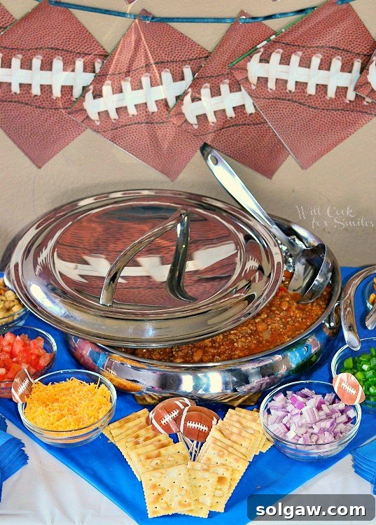 A large stainless steel bowl brimming with warm chili, topped with a lid and a silver spoon. The bowl sits on a football-themed table, surrounded by crackers and various chili toppings, ready for a party.