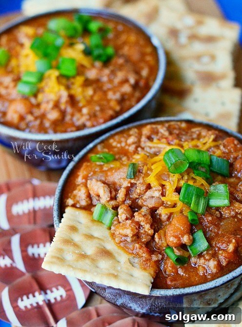 An overhead view of two small bowls of boozy chili, each generously topped with shredded cheese and fresh chives, presented on a vibrant blue platter alongside scattered crackers.