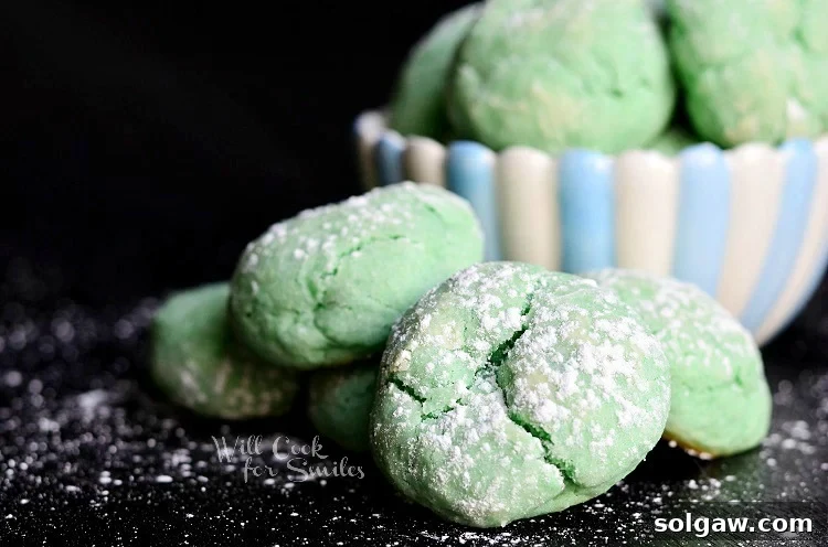 Cotton Candy Cloud Cookies 3 green Cotton Candy Cookies on a black counter top with powdered sugar and a bowl of white and blue cookies