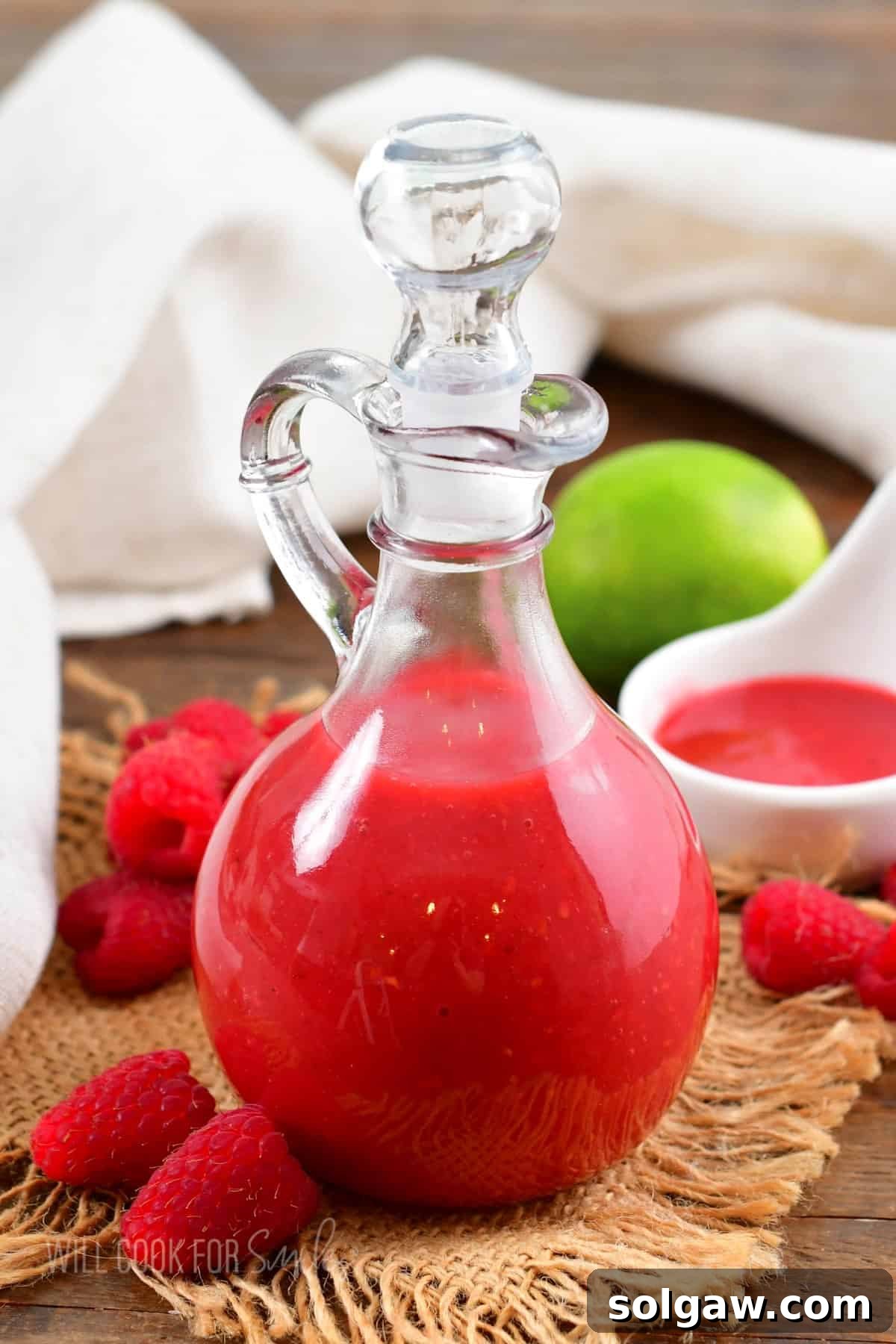 Sweet & Tangy Raspberry Drizzle 2 A tall glass dressing jar filled with bright pink homemade raspberry vinaigrette, surrounded by fresh raspberries and lime slices.