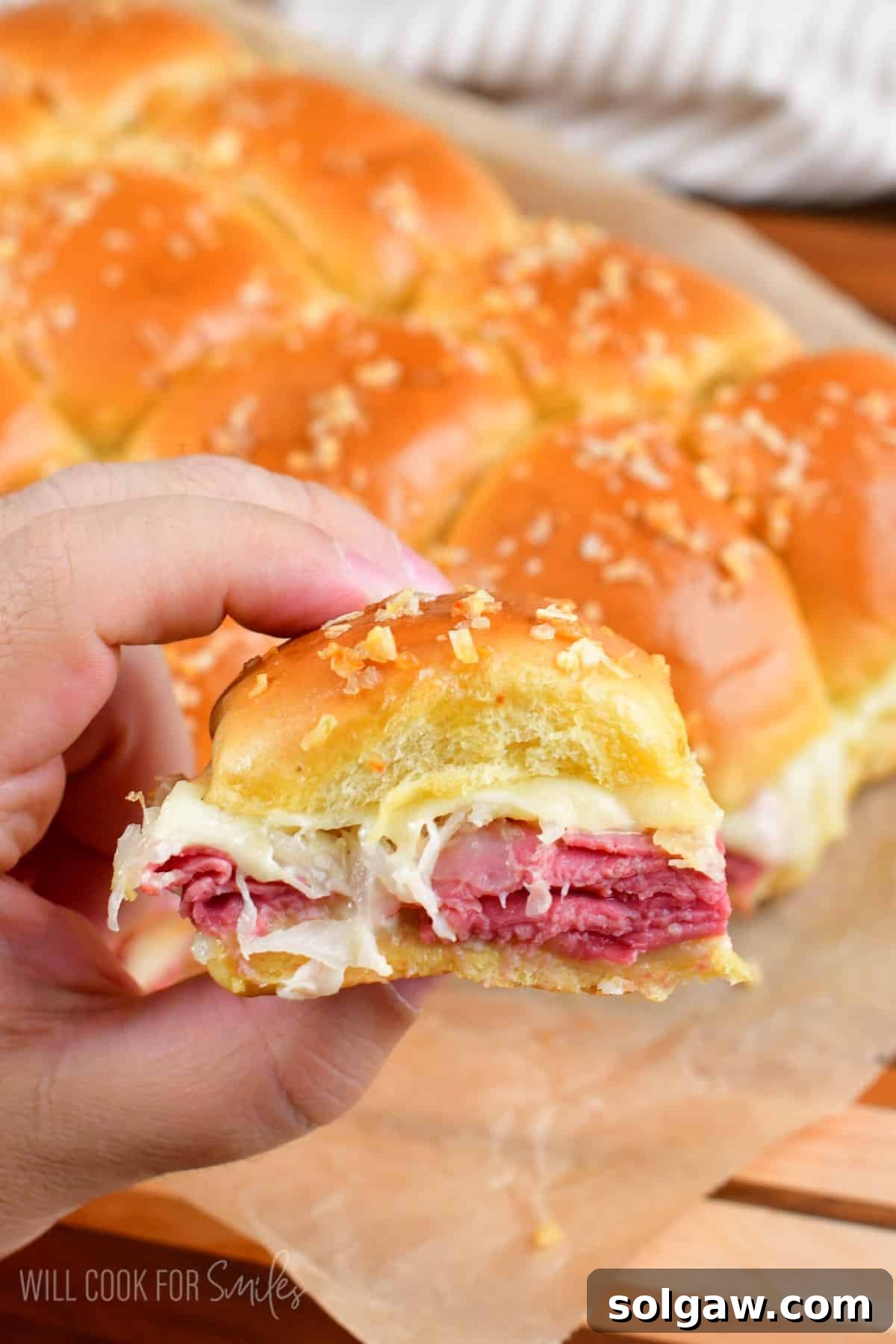 A hand holding a single Reuben slider, with the rest of the baked sliders blurred in the background, showcasing the melty cheese and golden bun.