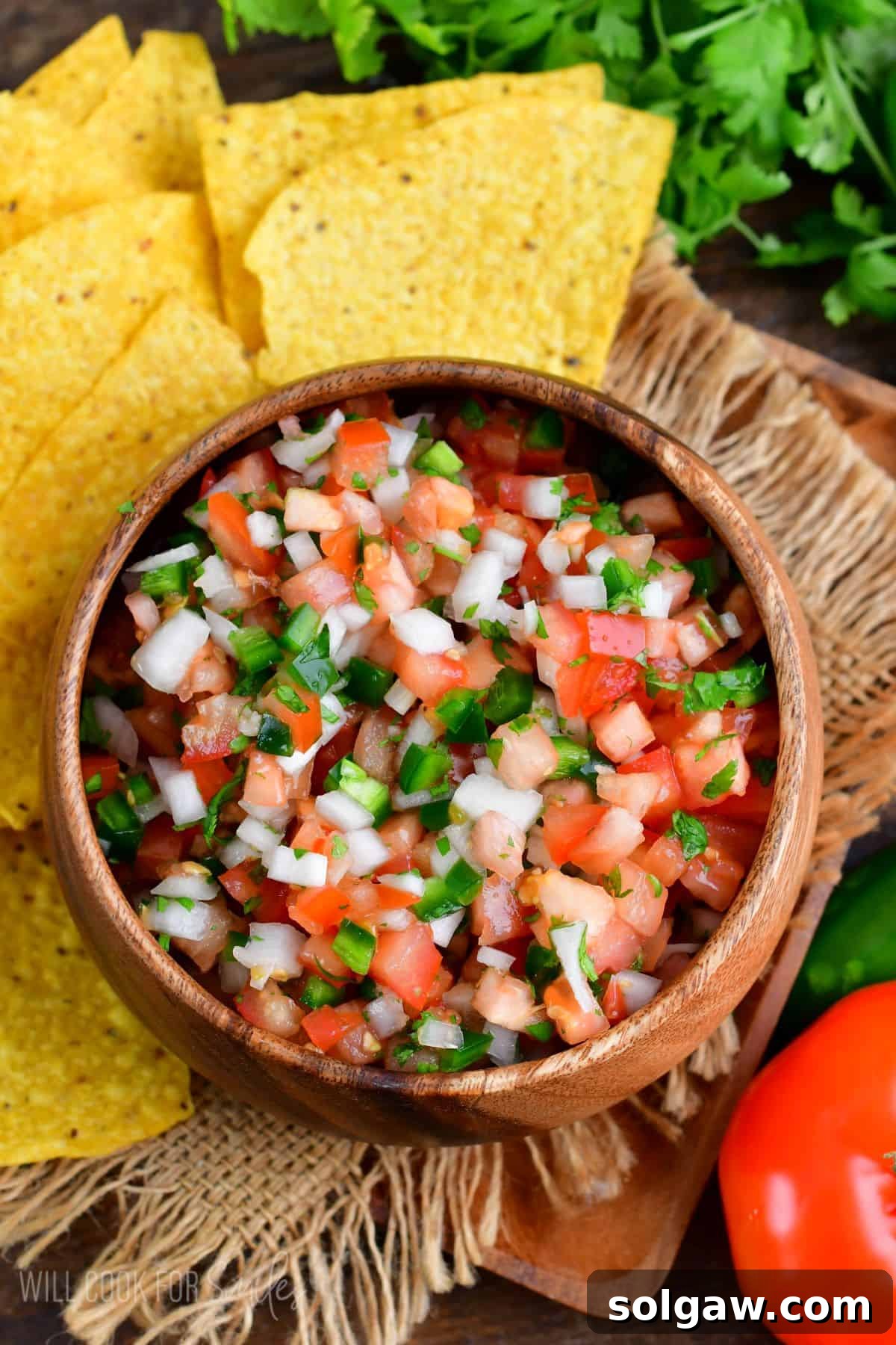 A rustic wooden bowl brimming with fresh Pico de Gallo, with crispy tortilla chips artfully arranged beside it.