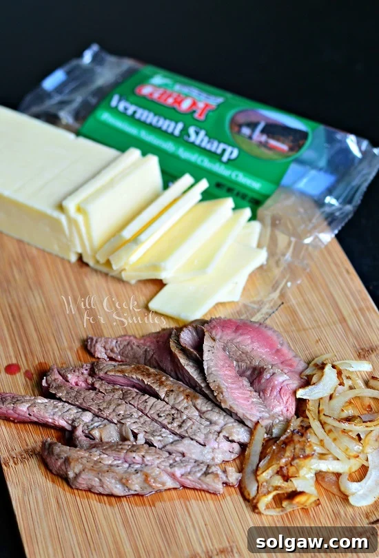 Sliced white cheddar cheese, cooked steak, and golden caramelized onions prepared on a cutting board, ready to be assembled into a grilled cheese.