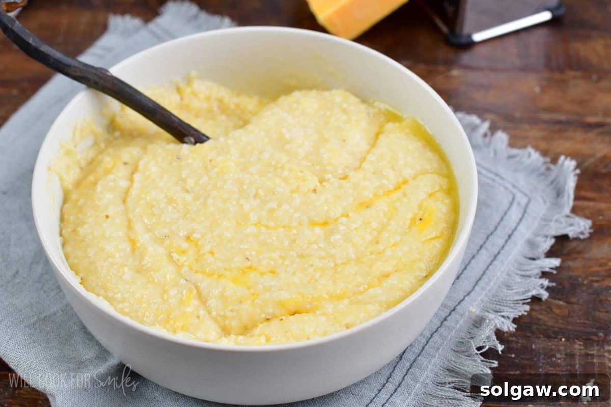 A close-up of a spoon stirring smooth, buttery, and cheesy grits in a white bowl, highlighting the creamy texture.