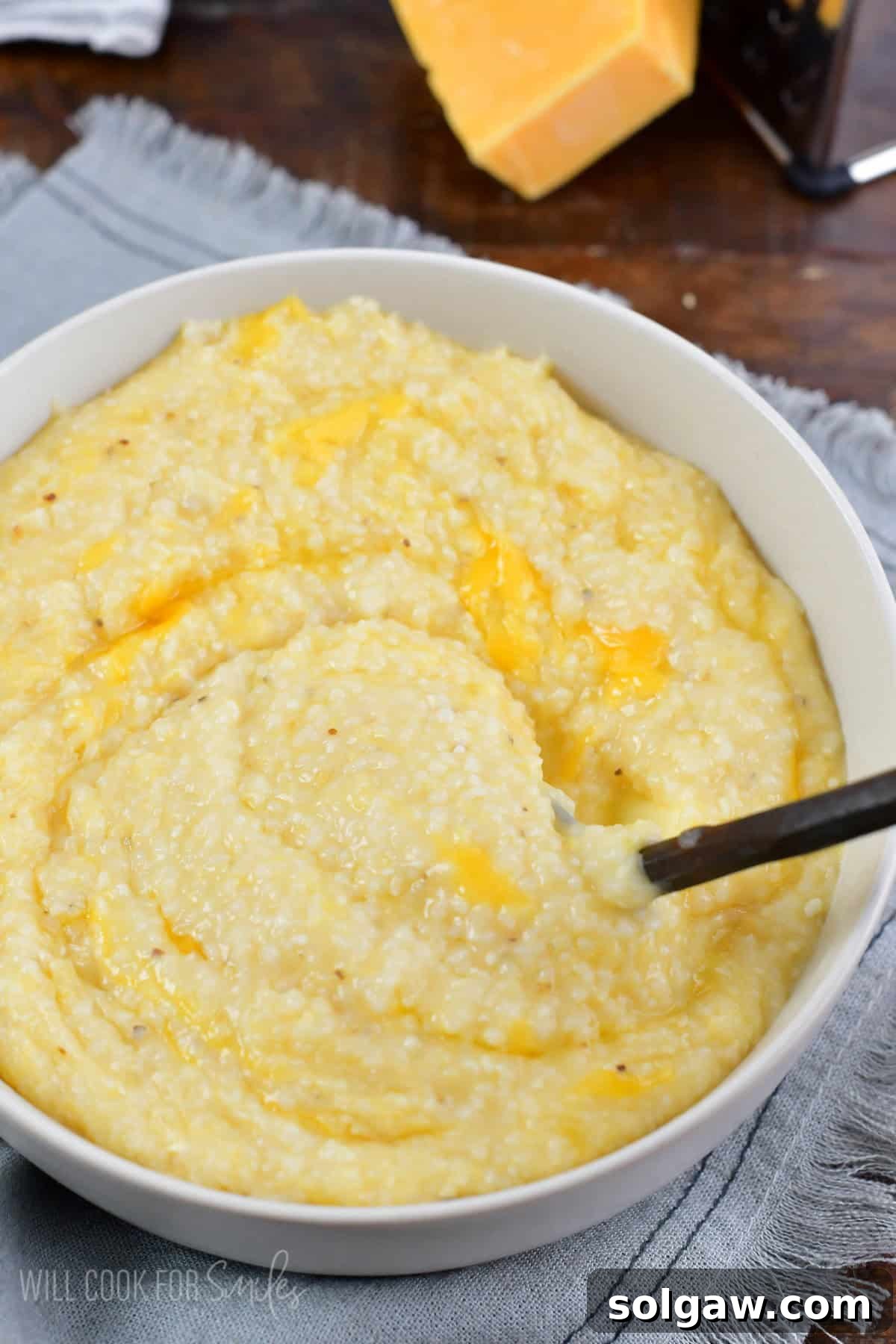 A hand stirring a bowl of rich, yellow cheese grits with a metal spoon, showcasing the smooth and thick texture.