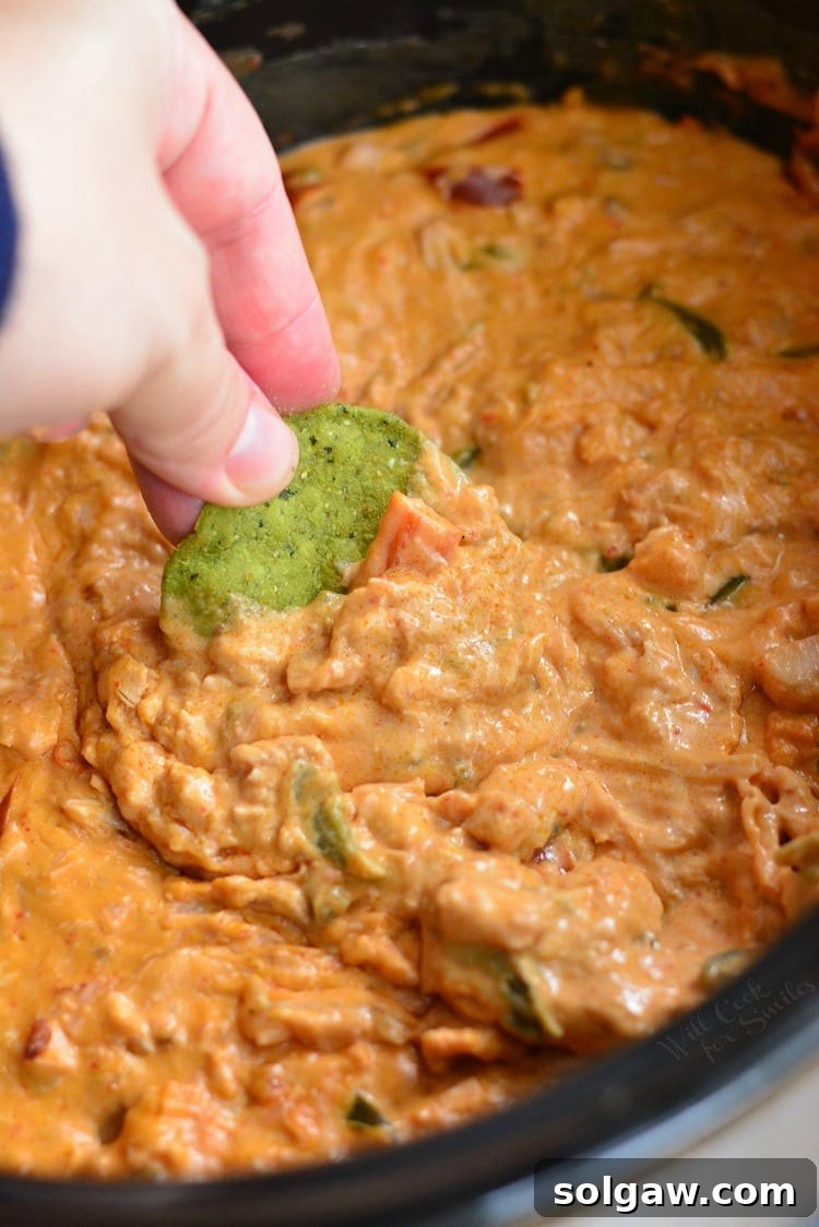 A green tortilla chip being dipped into the slow cooker enchilada dip, with melted cheese visible.