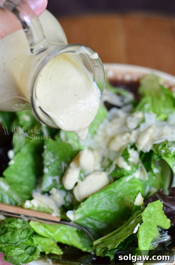Light homemade Caesar dressing being poured from a glass cruet onto a fresh, crisp Caesar salad in a white bowl, garnished with croutons.