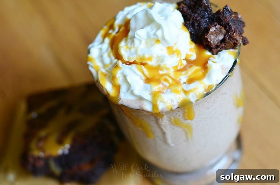 view from above of Glass mug filled with salted caramel brownie milkshake on a wooden table with a brownie in front of glass while both are drizzled with caramel