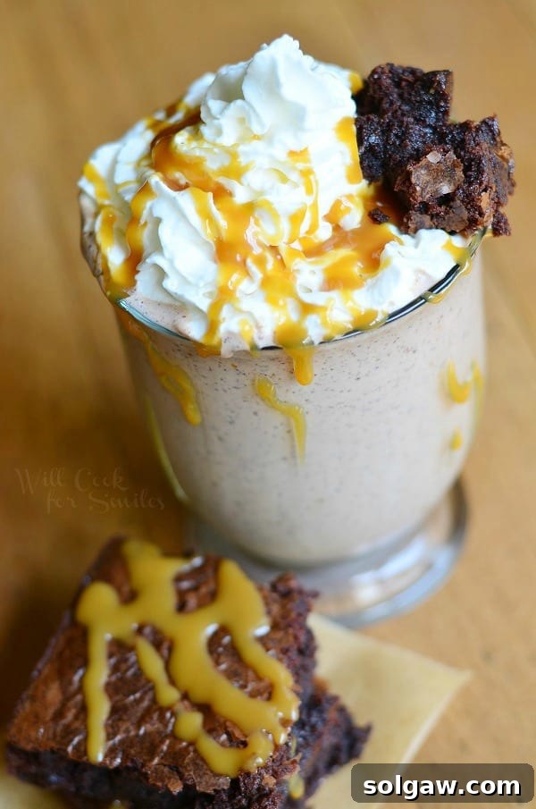 Salted Caramel Brownie Milkshake in a glass with whipped cream and caramel drizzle, a brownie piece on top, and another brownie next to the glass.