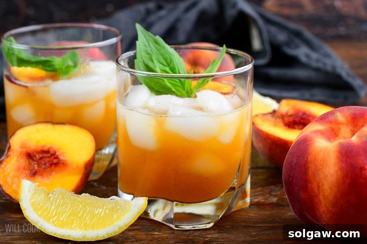 two glasses of peach smash bourbon in a glass with ice and peaches and mint on top as garnish on a wood surface.