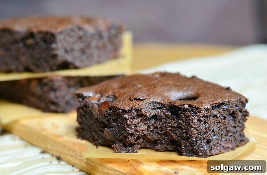 close up view of 2 dark chocolate chocolate chip brownies stacked with wax paper in between each stacked in background with 1 brownie in foreground on a wooden cutting board on a tand and white placemat on wood table