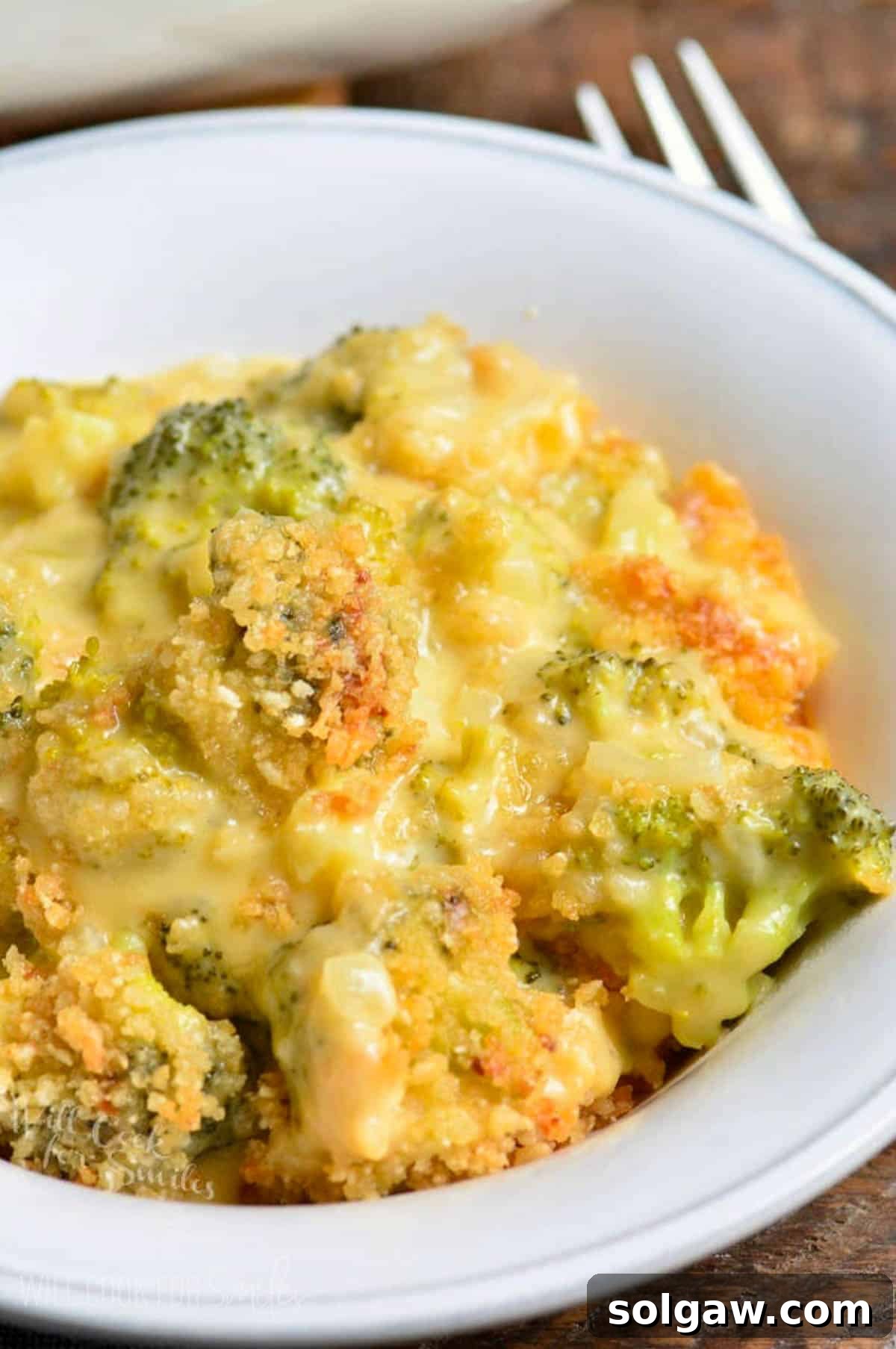 A close-up shot of a cheesy broccoli cheese casserole served in a white bowl, with a fork ready to scoop, highlighting its creamy texture.