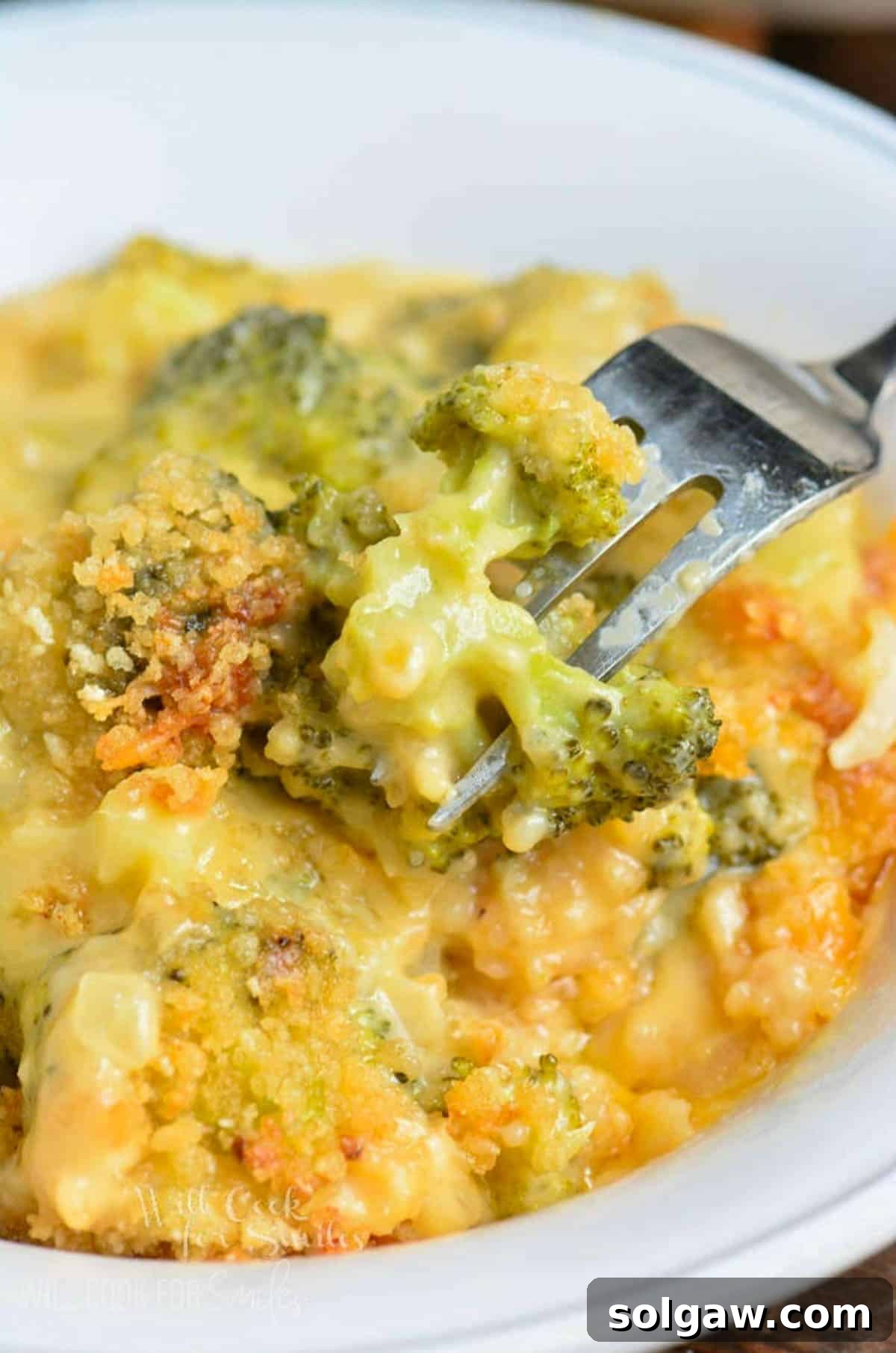A fork scooping a portion of cheesy broccoli cheese casserole from a bowl, highlighting its gooey texture.