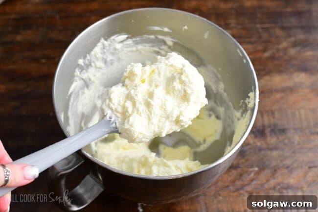 A spatula scoops a dollop of light and smooth white chocolate mousse frosting from a mixing bowl, showcasing its creamy texture.