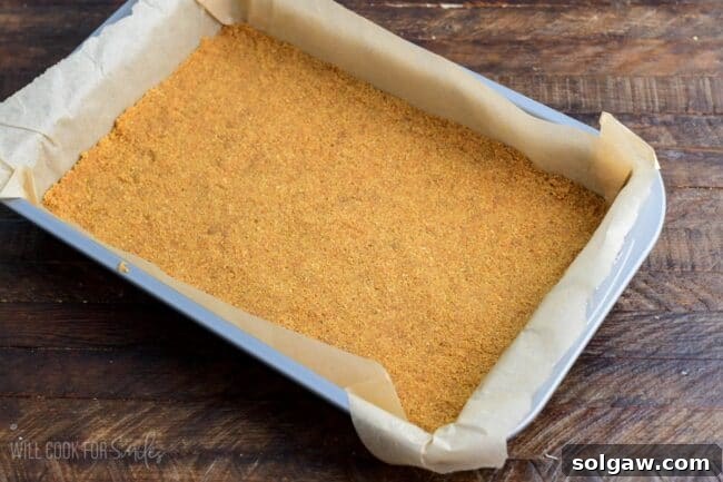 The perfectly pressed graham cracker crust for Christmas Cheesecake in a parchment-lined baking dish, awaiting the next layers, on a wooden surface.