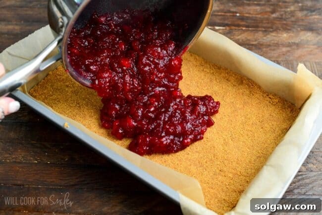 The vibrant cranberry jam being meticulously spread over the graham cracker crust in the baking pan, creating a beautiful base layer for the Christmas Cheesecake.