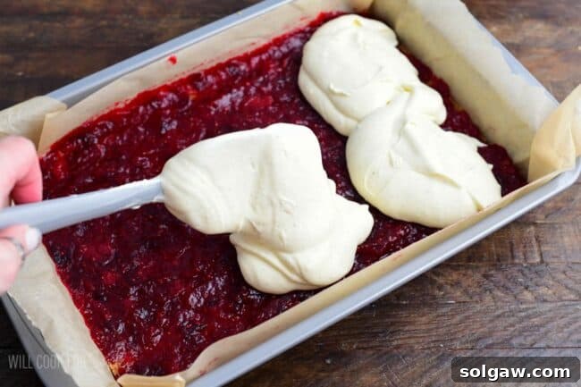 The creamy cheesecake filling being carefully spread over the vibrant cranberry sauce layer in the baking pan, preparing the Christmas Cheesecake bars for baking.