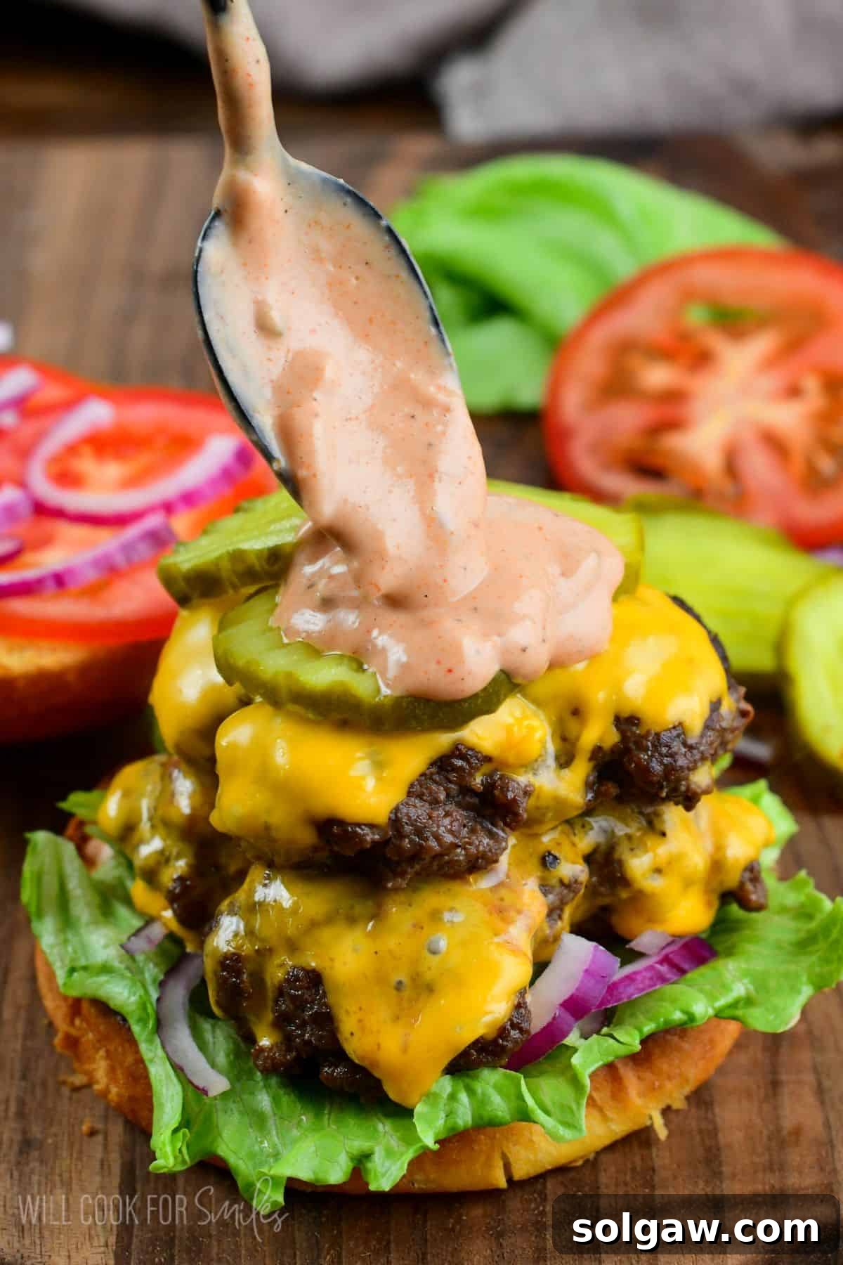 adding creamy burger sauce with a spoon on top of a juicy smashed burger with lettuce, pickle, onion, and cheese.