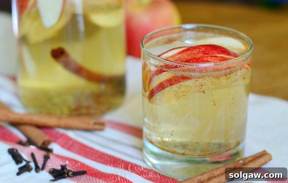 Close up view of small glass filled with spiced apple sangria with sliced apples floating near the top. A pitcher of sangria and apple are in the background.