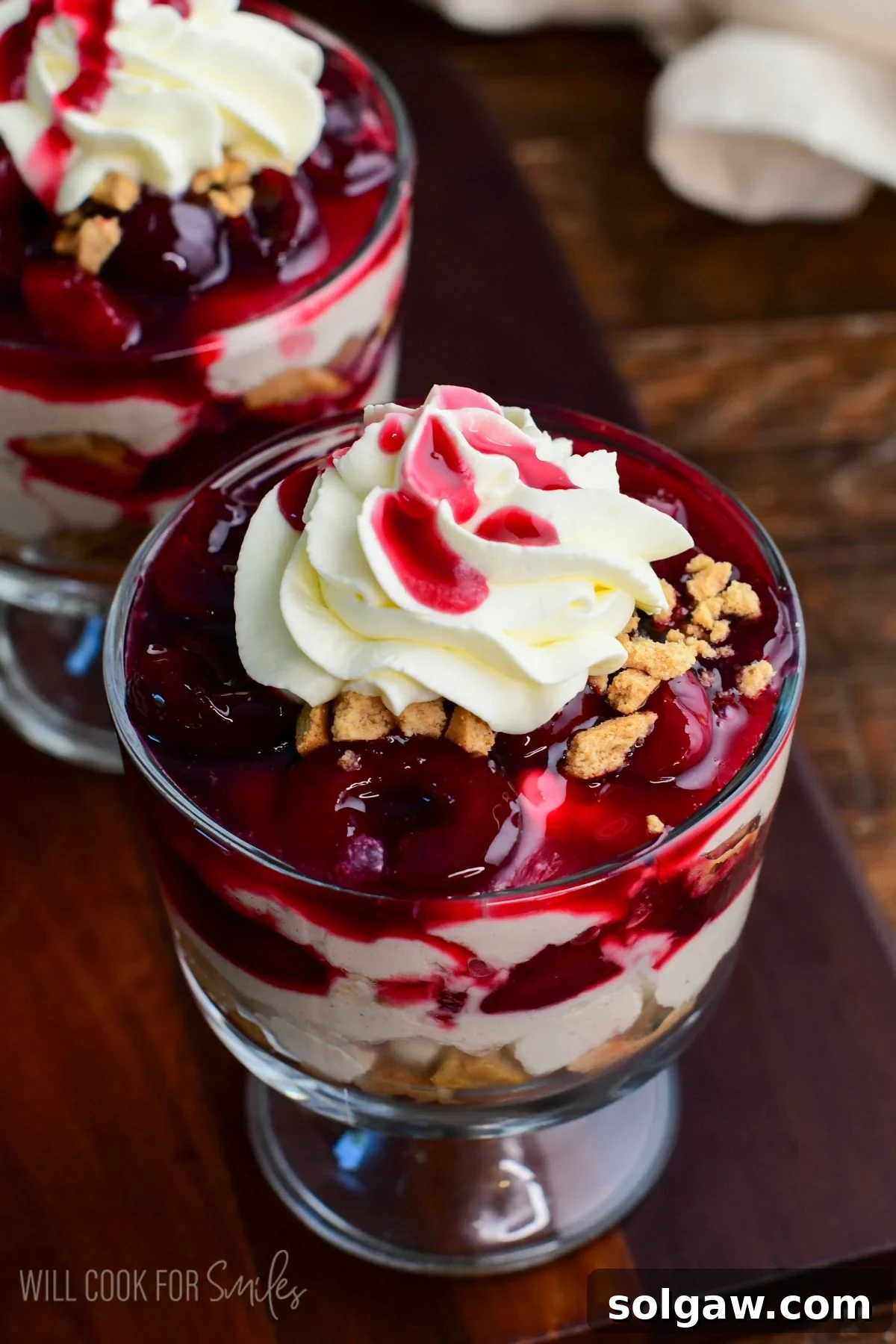 A stunning no-bake cherry cheesecake parfait, garnished with whipped cream, a drizzle of cherry sauce, and cookie crumbles, presented in a sleek glass dessert cup.