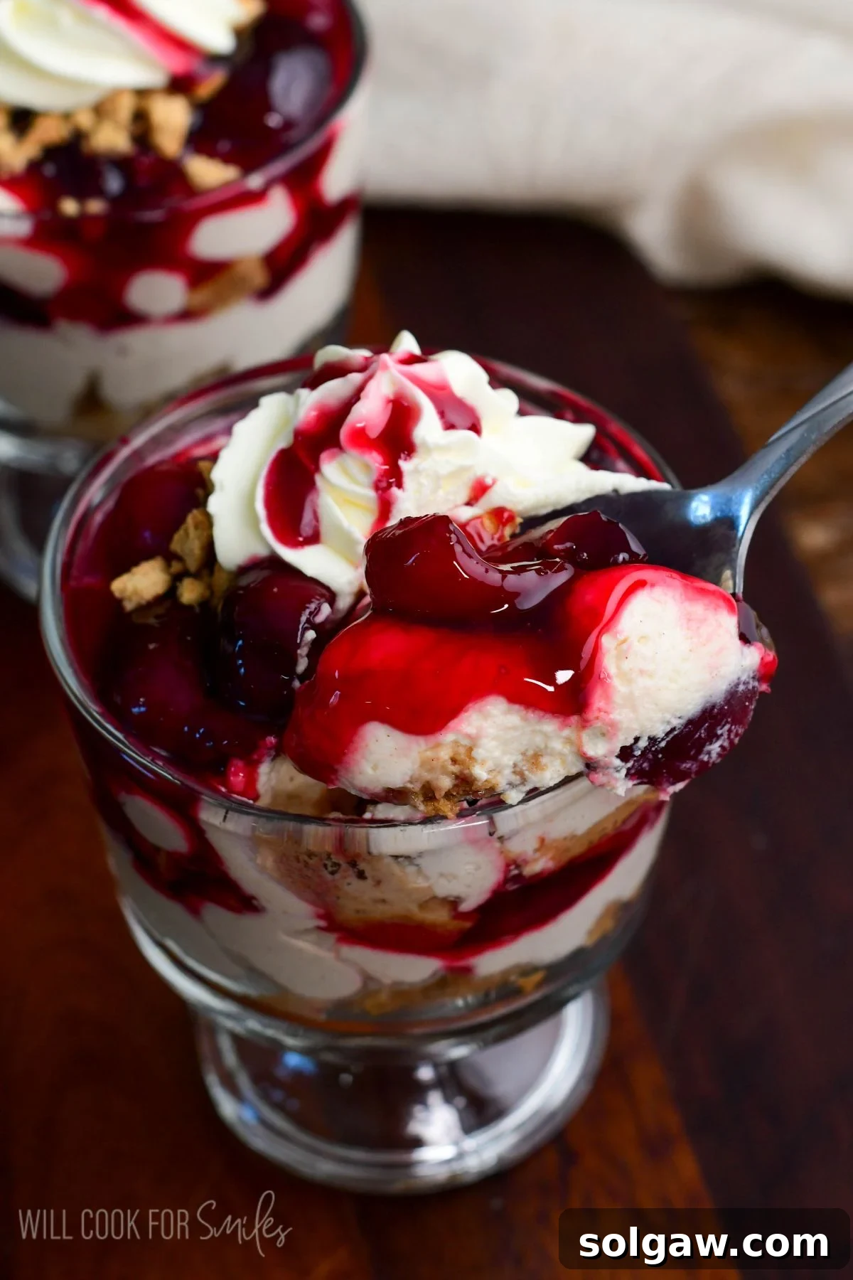 A spoon gracefully scoops into a no-bake cherry cheesecake parfait, revealing the creamy, fruity layers topped with whipped cream and cherry sauce.