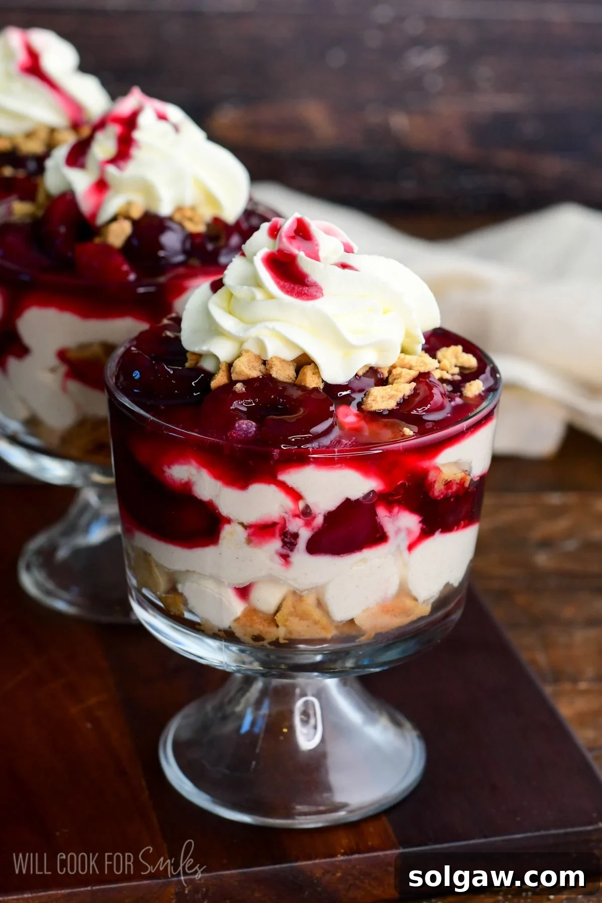 A close-up of a single no-bake cherry cheesecake parfait, featuring distinct layers of crushed cookies, creamy filling, and cherry topping in a glass, with a dollop of whipped cream on top.