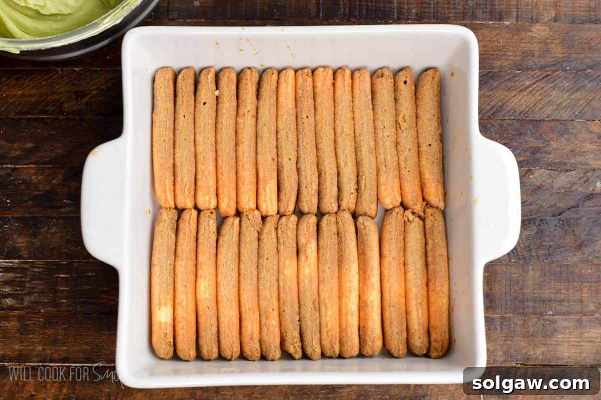 Velvet Pistachio Tiramisu 11 Layer of soaked ladyfinger cookies arranged neatly in a white baking dish, ready for pistachio tiramisu assembly.