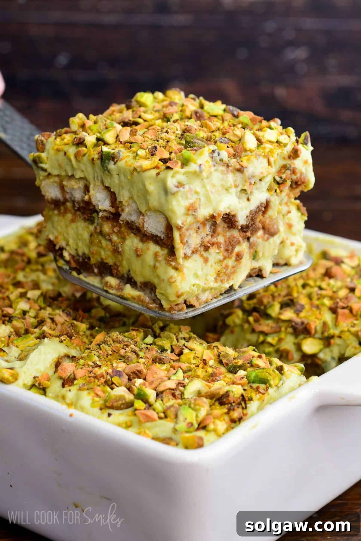 Velvet Pistachio Tiramisu 16 Close-up of a thick slice of pistachio tiramisu being lifted from a white baking dish, showing creamy pistachio layers, soaked ladyfingers, and a crunchy chopped pistachio topping.