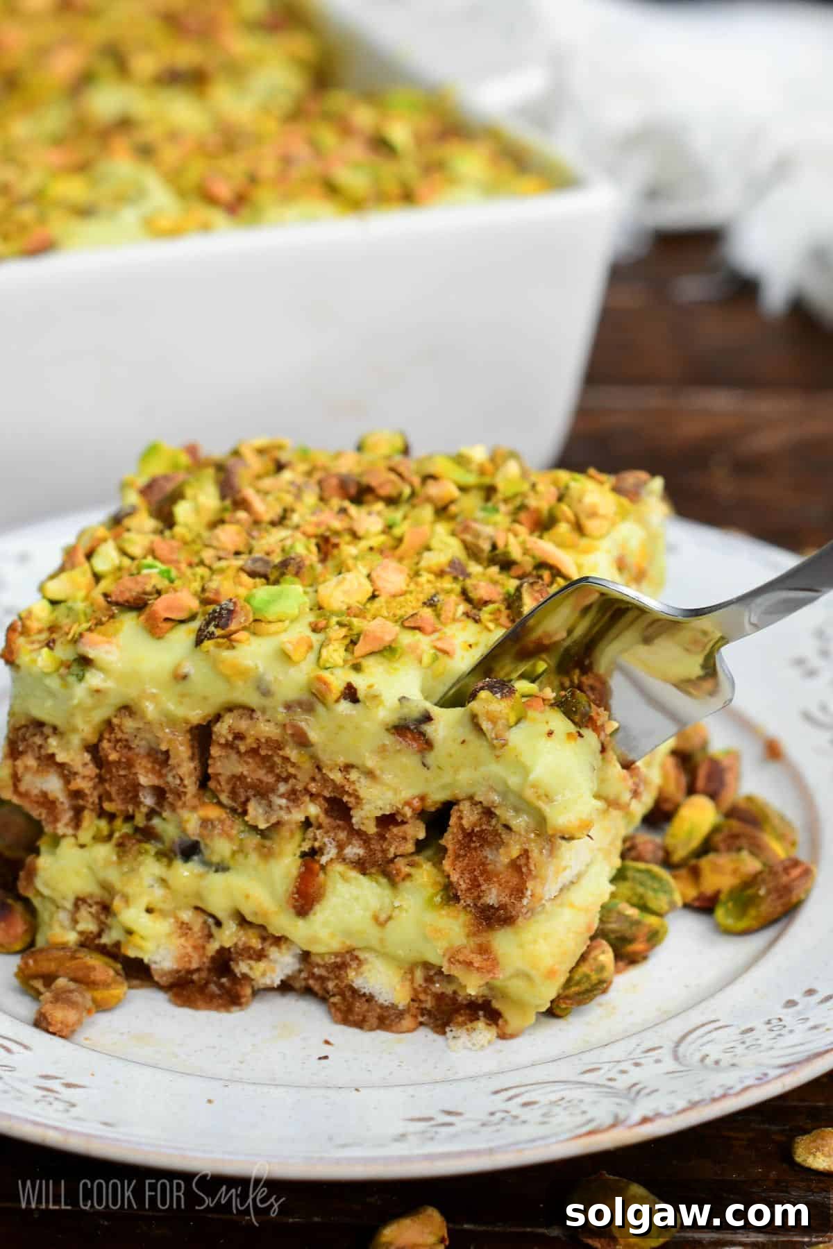 Velvet Pistachio Tiramisu 17 Fork cutting into a creamy slice of pistachio tiramisu topped with chopped pistachios, showing layers of pistachio mascarpone filling and coffee-soaked ladyfingers on a plate.