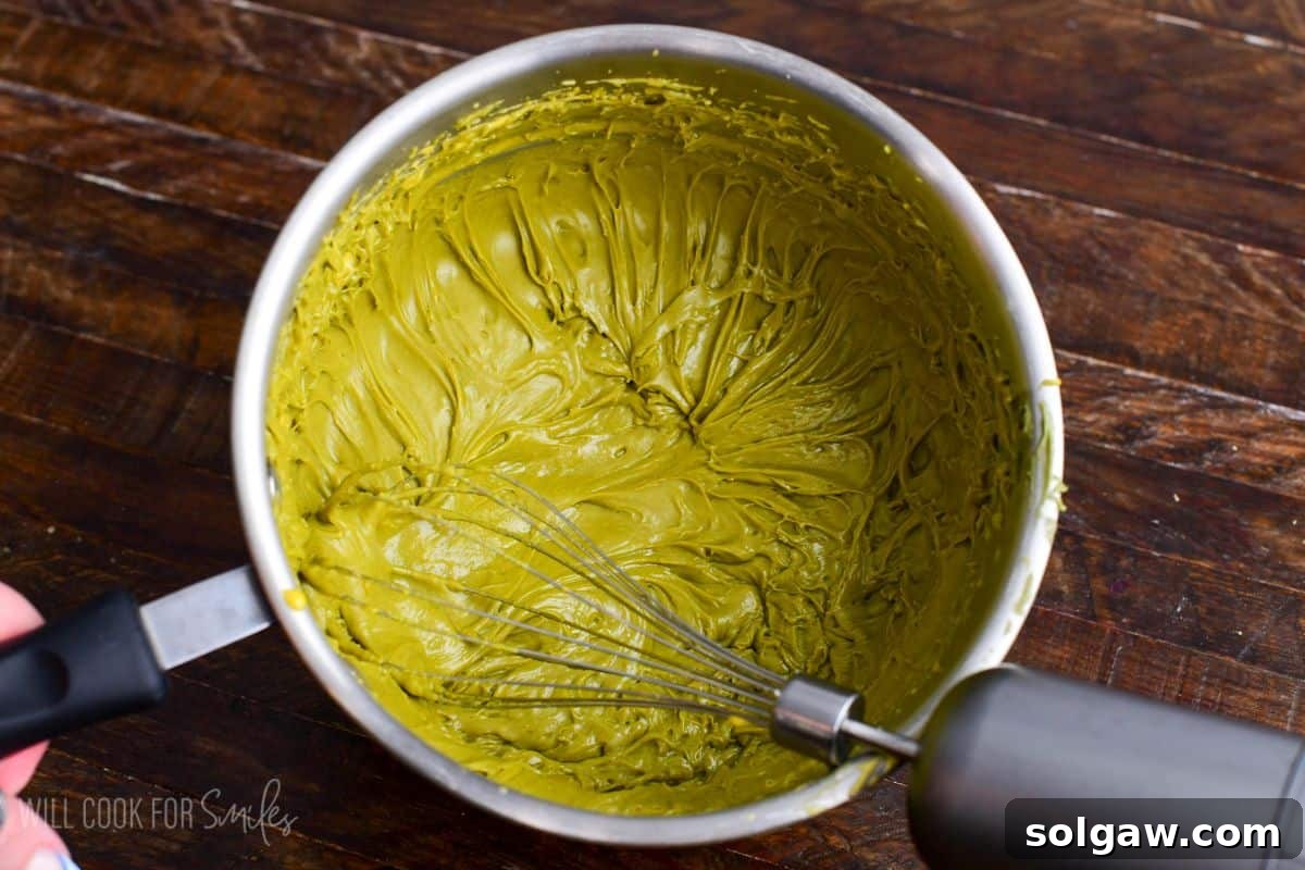Velvet Pistachio Tiramisu 8 Pistachio paste whisked into egg yolk custard in a saucepan, creating a thick green pistachio cream mixture.