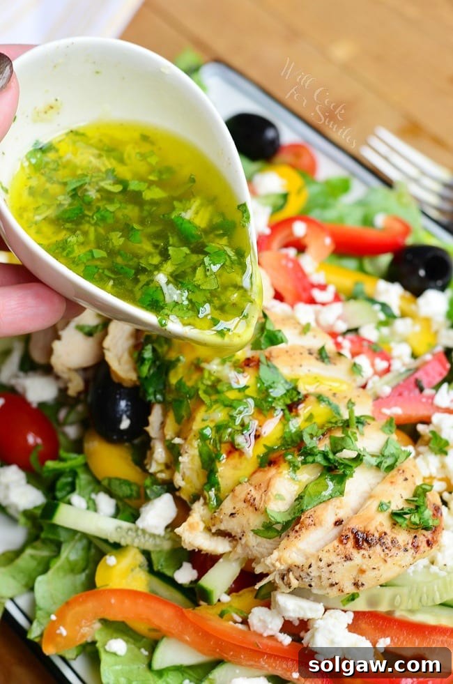 Pouring homemade lemon herb vinaigrette over freshly sliced chicken on a bed of Greek salad, highlighting the vibrant dressing.