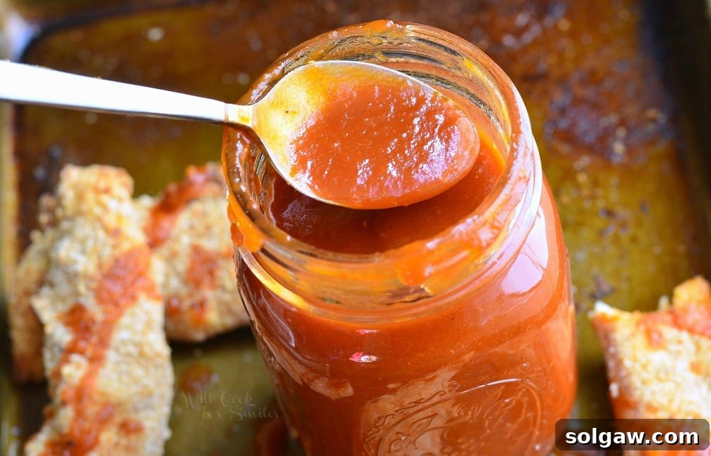 A close-up of Homemade Whiskey BBQ Sauce in a mason jar, with a spoon scooping out some sauce, next to a baking sheet of Easy Baked Chicken Strips.