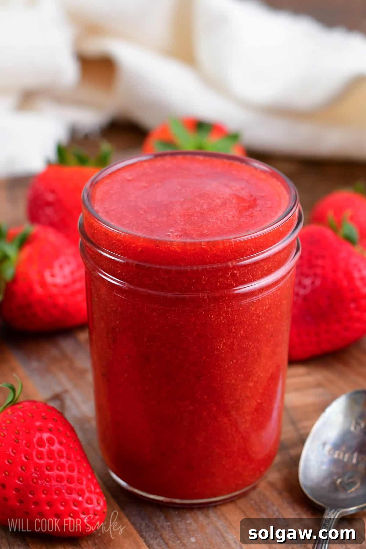 Freshly made strawberry sauce in a glass jar, surrounded by ripe strawberries on a rustic wooden surface.