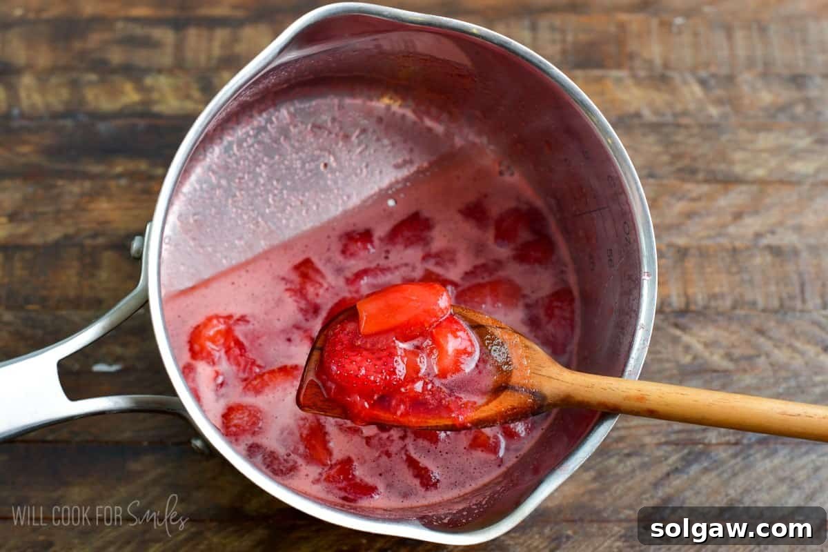 Cooked strawberries simmering in a pot, with some being gently scooped out with a wooden spoon to show the consistency.