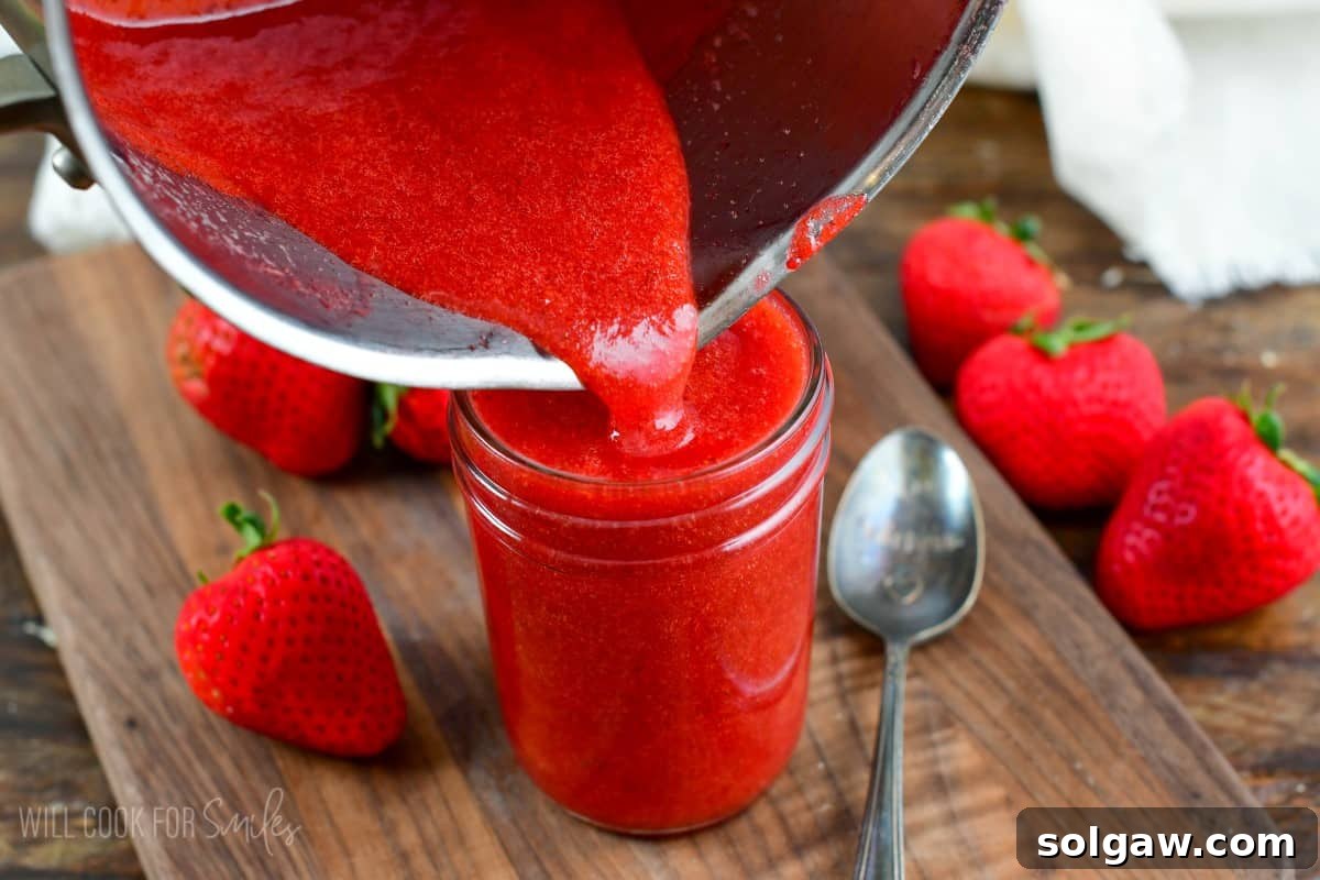 Pouring freshly made strawberry sauce from the pot into a clear mason jar on a wooden cutting board.