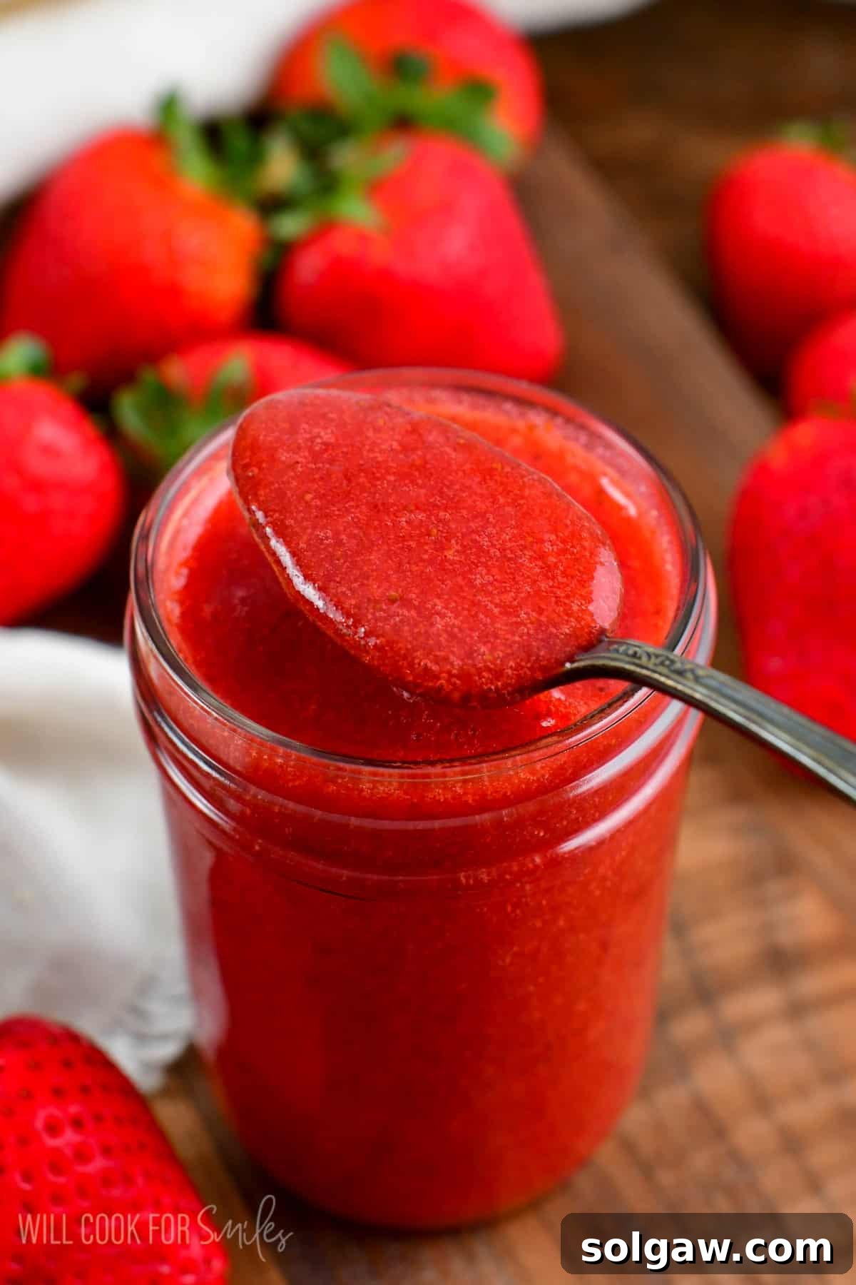 A glass jar of strawberry sauce with a spoon, surrounded by fresh strawberries on a light wooden surface.