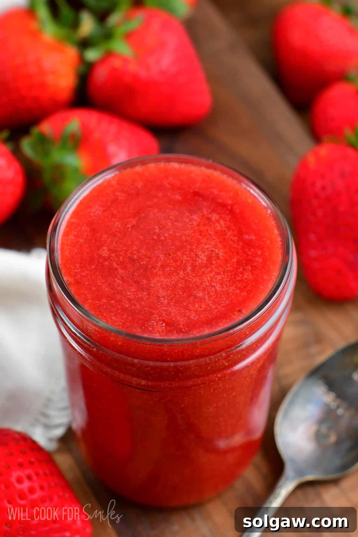 A mason jar filled with vibrant red strawberry sauce, a small spoon resting next to it, and fresh strawberries scattered around on a wooden surface.