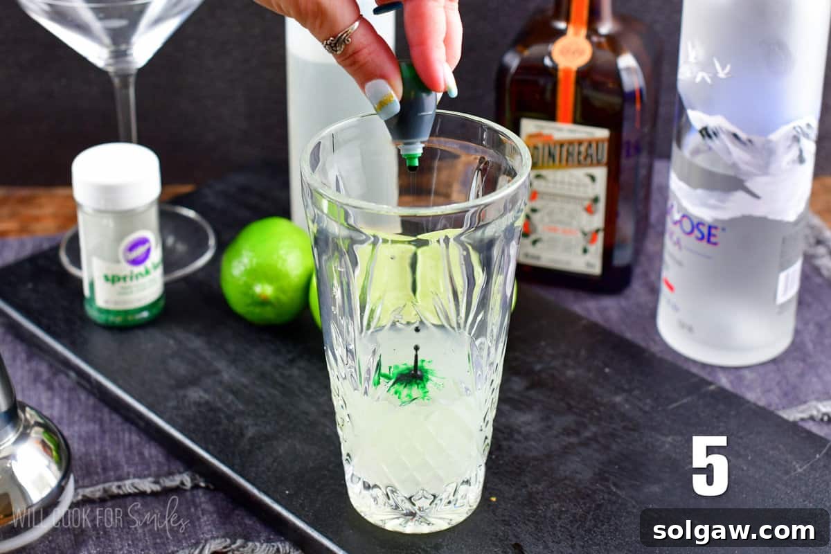 Two drops of vibrant green food coloring being added to the cocktail mixture in a glass shaker, with a bottle of vodka blurred in the background on a black wooden surface.