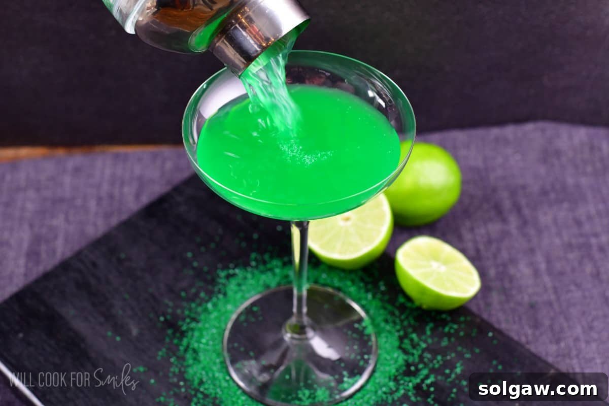 A vibrant green lime martini being poured from a shaker into an elegant martini glass, with green sprinkles adorning the base of the glass and a sliced lime ready for garnish.