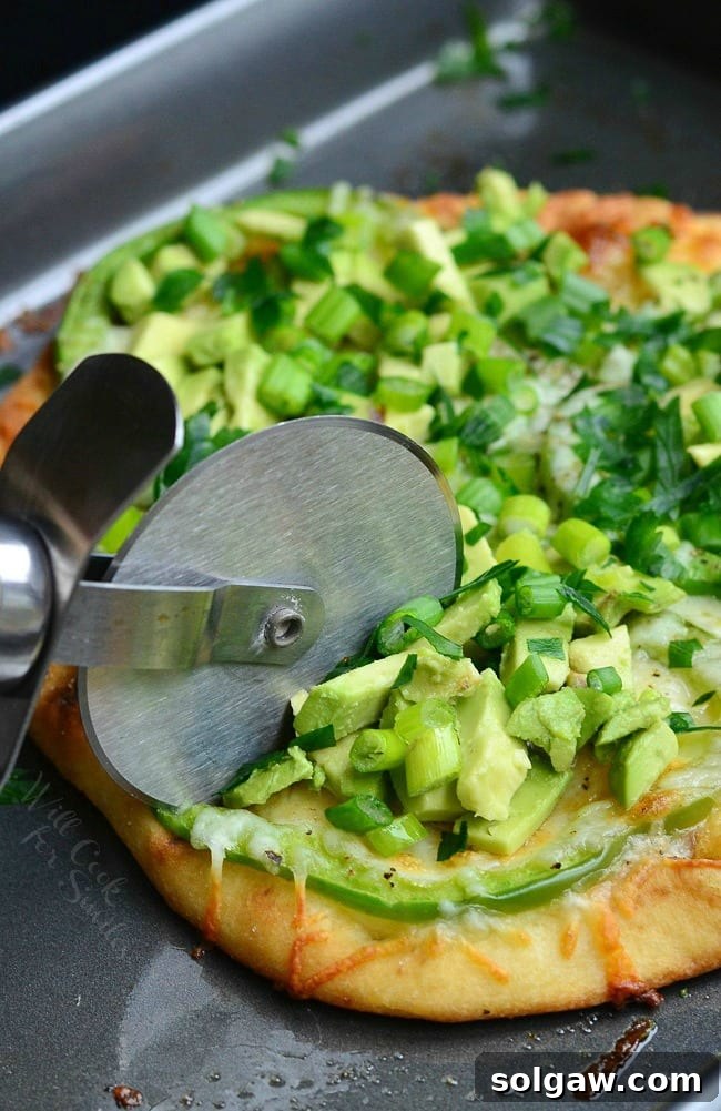 Slicing Green Naan Pizza with avocado, green peppers, and green onions on top using a pizza slicer.