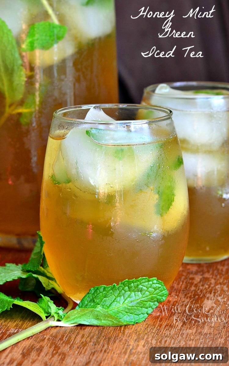 Two glasses of brewed Honey Mint Green Iced Tea on a wooden table with mint leaves, and a pitcher in the background.